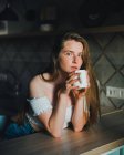 Спокойная молодая женщина с длинными волосами в повседневной одежде с голыми плечами наслаждается свежим горячим напитком и смотрит в камеру, опираясь на кухонный стол — стоковое фото