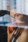 Mulher sonhadora atraente em top branco com ombros nus segurando pêssego doce e olhando para a câmera através do vidro — Fotografia de Stock