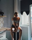 Unbewegte junge Frau in kurzen Hosen und Top mit nackten Schultern sitzt mit einer Tasse Getränk auf dem Küchentisch und blickt gelassen in die Kamera — Stockfoto