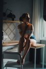 Unemotional young female in shorts and top with bare shoulders sitting on kitchen counter and looking away calmly — Stock Photo