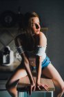 Unbewegte junge Frau in kurzen Hosen und Top mit nackten Schultern sitzt auf dem Küchentisch und schaut ruhig weg — Stockfoto