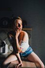 Unbewegte junge Frau in kurzen Hosen und Top mit nackten Schultern sitzt auf dem Küchentisch und schaut ruhig weg — Stockfoto