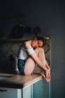 Неэмоциональная молодая женщина в шортах и топе с голыми плечами сидит на кухонном столе и спокойно смотрит в камеру — стоковое фото
