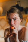 Mulher sonhadora atraente em top branco com ombros nus segurando pêssego doce e olhando para a câmera enquanto se inclina no balcão da cozinha — Fotografia de Stock