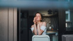 Unemotional young female with top with bare shoulders sitting on kitchen counter and looking at camera calmly — Stock Photo