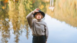 From above young blond cool female tourist in trendy apparel looking at camera while standing on coast against water reflecting trees — Stock Photo