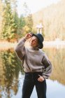 Молода крута жінка-туристка в модному одязі, дивлячись геть, стоячи з рукою в кишені на узбережжі проти води, що відображає дерева — стокове фото