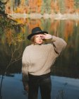 Joven turista rubia fresca con ropa de moda mirando a la cámara mientras está de pie cubriendo la cara en la costa contra los árboles reflejantes de agua - foto de stock