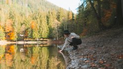 Seitenansicht einer Reisenden in lässiger Kleidung und Hut, die sich im transparenten Wasser gegen Bäume im Herbst spiegelt — Stockfoto