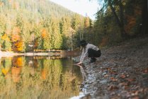Вид сбоку анонимной женщины-путешественницы в повседневной одежде и шляпе, отражающейся в прозрачной воде против деревьев осенью — стоковое фото