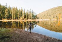Back view of anonymous female tourist in hat with raised arms admiring transparent water against autumn trees on mount — Stock Photo