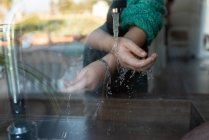 Através de vidro de colheita criança irreconhecível lavar as mãos sob água corrente em pia na cozinha — Fotografia de Stock