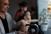 Маленькая девочка моет голову милого ребенка на руках отца во время купания в раковине на кухне — стоковое фото