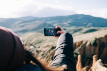 Back view of unrecognizable tourists in outerwear taking self portrait on cellphone while resting on mount against ravine in back lit — Stock Photo