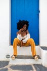 Joven mujer afroamericana en traje de verano de moda sentada en la puerta cerca del edificio en la vieja calle de la ciudad - foto de stock
