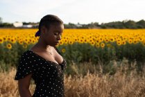 Vista lateral de la mujer afroamericana en vestido de pie sobre fondo de girasoles florecientes en el campo y disfrutando del verano en el campo - foto de stock