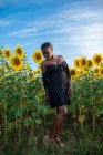 Vista lateral da fêmea afro-americana de vestido em pé no fundo de girassóis florescentes no campo e desfrutando de verão no campo — Fotografia de Stock