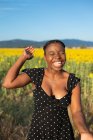 Mulher afro-americana alegre de vestido em pé no campo florescente com girassóis enquanto ri com os olhos fechados e desfruta de dia ensolarado no verão — Fotografia de Stock