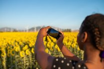 Visão traseira de uma mulher afro-americana irreconhecível tirando fotos do campo de girassol florescente no verão enquanto usa smartphone e passa o fim de semana no campo — Fotografia de Stock