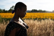 Вид сбоку афроамериканки в платье, стоящей на фоне кусающихся подсолнухов в поле и наслаждающейся летом в сельской местности — стоковое фото
