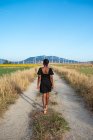 Vista trasera de la irreconocible mujer afroamericana en vestido de verano caminando por el camino rural en el campo sobre el fondo del campo de girasol y las montañas - foto de stock