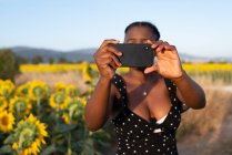 Афроамериканка фотографирует цветущее подсолнечное поле летом, пользуясь смартфоном и проводя выходные в сельской местности — стоковое фото