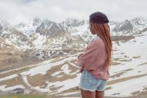 Seitenansicht einer nicht wiedererkennbaren Afroamerikanerin in stylischem Outfit, die im Winter vor dem Hintergrund schneebedeckter Berge steht — Stockfoto