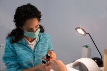 Attentive female manicurist in face mask applying lacquer on nail of crop woman near shiny lamp in beauty salon — Stock Photo