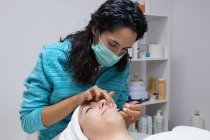 Анонимный косметолог в маске, лечит взрослую женщину с закрытыми глазами во время процедуры по уходу за лицом в салоне красоты — стоковое фото
