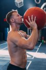 Vista lateral del deportista con torso desnudo lanzando balón medicinal durante el entrenamiento funcional en el gimnasio - foto de stock