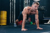 Atleta masculino enfocado con torso desnudo muscular haciendo flexiones durante el entrenamiento funcional y mirando hacia adelante - foto de stock