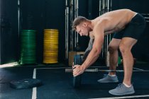 Side view of muscular sportsman with strong naked body putting heavy weight plate on barbell while preparing for functional training in gym — Stock Photo