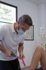 Анонимный внимательный физиотерапевт-мужчина в стерильной маске перерезает эластичную кинезиологическую ленту ножницами в больнице — стоковое фото