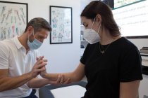 Анонимный хиропрактик-мужчина в респираторной маске изучает предплечье женщины в процессе физиотерапии в медицинском центре — стоковое фото