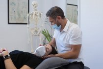 Неузнаваемый физиотерапевт-мужчина в маске для лица проверяет шею женщины возле человеческого скелета в медицинском центре — стоковое фото