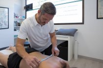 Fisioterapeuta masculino masajeando la espalda de una mujer anónima en topless con herramienta de acero inoxidable en la clínica - foto de stock