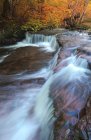 Long exposure picturesque scenery of shallow rapid stream flowing among rough boulders between deciduous trees on autumn day — Stock Photo