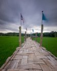 Long pont en bambou Su Tong Pae avec divers drapeaux sur des piliers en bois traversant un champ de riz contre un ciel nuageux en Thaïlande — Photo de stock
