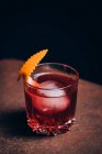 Copa de refrescante cóctel Negroni con sabor amargo y hielo adornado con cáscara de naranja y servido en el brazo del sofá en habitación oscura — Stock Photo