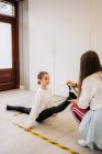 Side view of flexible girl sitting in split and stretching legs with help of dance trainer before lesson in spacious hall — Stock Photo