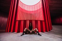 Artista de teatro étnico sentado com as pernas afastadas no chão perto de parede com nervuras brilhantes no palco e olhando para a câmera — Fotografia de Stock