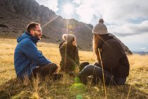 Viajeros alegres en ropa de abrigo hablando mientras descansan en la hierba contra la cresta bajo el cielo nublado a la luz del sol - foto de stock