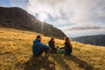 Веселые путешественники в верхней одежде разговаривают во время отдыха на траве против хребта под облачным небом под солнечным светом — стоковое фото