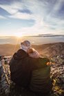 Back view of unrecognizable man embracing girlfriend in outerwear while admiring dawn from mountain under cloudy sky in sunshine — Stock Photo