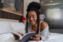 Счастливая молодая афроамериканская студентка в повседневной одежде лежит на удобной кровати и читает книгу дома — стоковое фото
