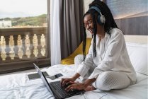 Vista lateral de una joven estudiante afroamericana positiva con ropa casual y auriculares sonriendo mientras está sentada en el acogedor be y escribiendo en la computadora portátil durante la lección en línea en casa - foto de stock