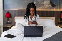 Concentrada joven afroamericana freelancer con pelo largo rizado en ropa casual y anteojos sentados en cómoda cama con portátil y portátil y el uso de teléfono inteligente durante el trabajo remoto - foto de stock