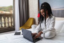 Konzentrierte junge afroamerikanische Freiberuflerin mit lockigem langen Haar in lässiger Kleidung und Brille sitzt während der Fernarbeit auf einem bequemen Bett mit Laptop und Notebook — Stockfoto