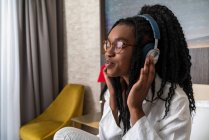 Vista lateral de la joven mujer afroamericana positiva en ropa casual y auriculares sonriendo con los ojos cerrados sentado en la acogedora cama con teléfono inteligente en casa - foto de stock