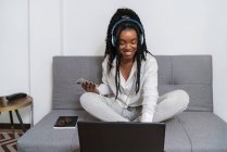 Joven dama afroamericana alegre con el pelo largo rizado en ropa casual sonriendo mientras escucha música en auriculares con teléfono inteligente y trabajando en la computadora sentada en el sofá en casa - foto de stock